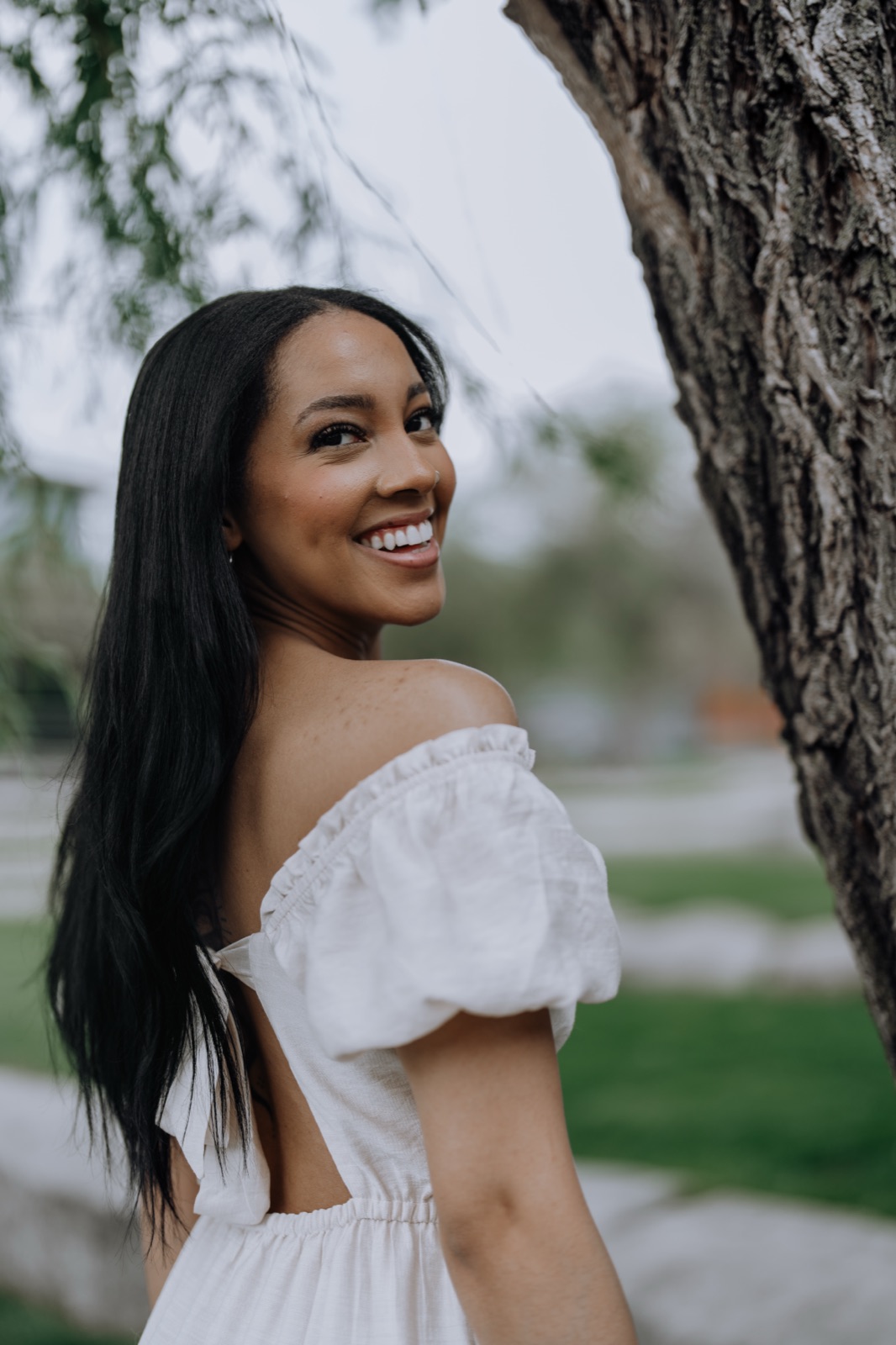 Portrait of a woman in a white dress, smiling over her shoulder beside a tree